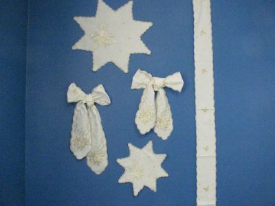 A blue wall with decorative white fabric items displayed: two star-shaped pieces, two bow-shaped pieces with candlelight embroidery, and a vertical white strip with a scalloped edge and white & gold embroidery designs.