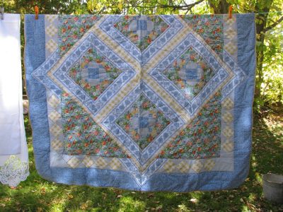 The 7-pieces French Country Quilt & Sheet Bundle Deal with a floral pattern hangs on a clothesline outdoors. It showcases diamond-shaped designs in blue, yellow, and green, surrounded by trees and grass.