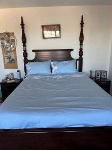 A neatly made bed, featuring the 7-pieces French Country Quilt & Sheet Bundle Deal in lsky blue with matching pillows, is centered between two wooden bedside tables. A framed picture hangs above, while white walls and mixed decorations adorn the side tables.