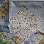A sky blue embroidered cloth with a delicate white lace edging rests on a 7-piece French Country Quilt, featuring colorful floral and checkered patterns. A tag is attached to the lace piece about the sheet, enhancing the vibrant backdrop.