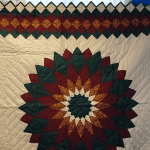The Nostalgia Brand 1980s Giant Dahlia Handmade Quilt features a central star pattern in green, burgundy, and yellow with intricate diamond borders against a cream background, displayed on a blue wall.