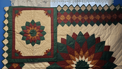 The Nostalgia Brand 1980s Giant Dahlia Handmade Quilt hangs showcasing bold star-like patterns in red, green, and gold. It includes intricate geometric designs and a border of alternating diamonds, giving it a traditional and cozy look. A sham is visible.