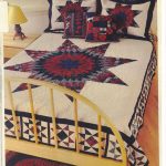 A bed with a wooden frame is adorned with the Flannel Tartan Lone Star hand quilted quilt, which features a large red, blue, and Cream star pattern. Matching pillows along with a small nightstand holding a yellow lamp are nearby, enhancing the cozy bedroom ambiance.