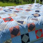 A MUSEUM FLANNEL PLAID PATCHWORK QUILT with ticking fabric backing is draped over a bed outdoors, showcasing its geometric pattern comprised of blue, red, black, and beige squares and triangles. Sunlight softly highlights the textures and colors against the backdrop of green grass.