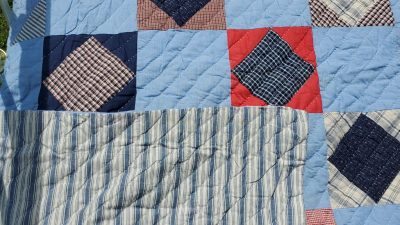 A quilt named "Museum Flannel Plaid Patchwork Quilt Ticking Fabric Backing" showcases a patchwork design with blue, red, and plaid squares. Each square presents a variety of fabric textures and colors, while a striped ticking pattern showcasing the backside fabric of the quilt.