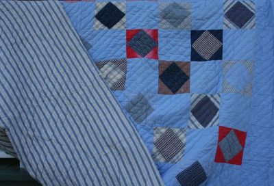 The MUSEUM FLANNEL PLAID PATCHWORK QUILT features an array of colorful patterns, such as plaid and solid squares, in shades of blue, red, and white. One corner is pulled back to reveal the ticking fabric backing with a striped design.