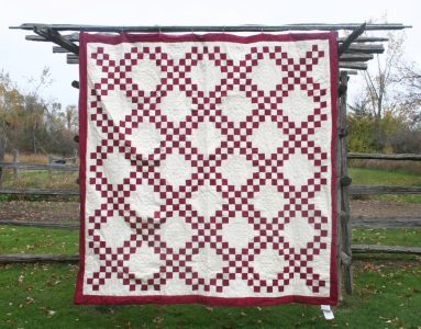 The Irish Chain Quilt–Burgundy RED, a handmade quilt with a red and ivory interlocking rings design, hangs outdoors on a wooden fence amid grass and autumn trees.