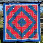 An Amish RED Barn Raising Handmade Flannel Quilt hanging outside on a wooden structure, showcasing a geometric pattern with vibrant red, blue, green, and gray squares arranged symmetrically. The quilt is displayed against a backdrop of grass and a clear blue sky.
