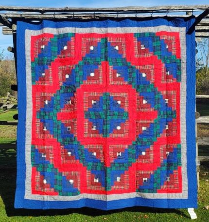 An Amish RED Barn Raising Handmade Flannel Quilt hanging outside on a wooden structure, showcasing a geometric pattern with vibrant red, blue, green, and gray squares arranged symmetrically. The quilt is displayed against a backdrop of grass and a clear blue sky.