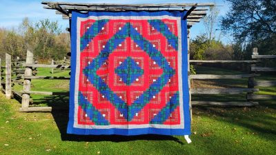 An Amish RED Barn Raising Handmade Flannel Quilt, with its vibrant geometric pattern of red, blue, and green squares forming a diamond shape at the center, hangs outdoors on a wooden structure. The serene setting is complemented by surrounding grass, trees, and a wooden fence.