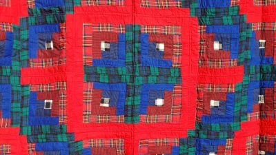 Introducing the Amish RED Barn Raising Handmade Flannel Quilt, characterized by its vibrant design of alternating squares in red, blue, and green plaid patterns. Each square showcases a textured, layered look that forms a symmetrical, geometric arrangement across the quilt.