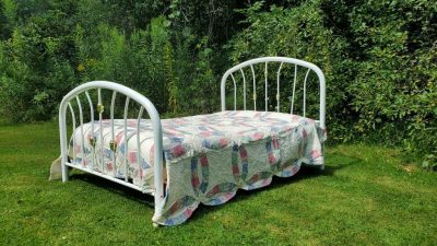 A white metal bed frame adorned with the Courtship Wedding Ring Orange Peel hand quilting is set up outdoors on a grassy lawn, surrounded by lush green foliage.