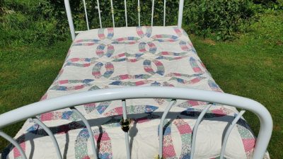 A white metal bed frame adorned with a Courtship Wedding Ring Orange Peel quilting design showcasing a colorful interlocking ring pattern is set on a grassy lawn, surrounded by lush green foliage.