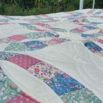 A close-up of the "Courtship Wedding Ring Orange Peel" quilt showcases a patchwork design with various floral patterns in pink, blue, green, and white. The quilt is spread out on a bed with a metal frame, and lush greenery can be seen in the background. Hand quilting featured Orange Peel design.