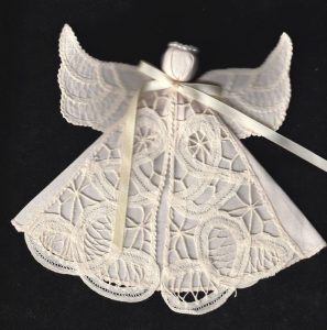 A lace angel ornament with a delicate embroidered pattern, featuring wings and a ribbon tied at the neck, set against a dark background.