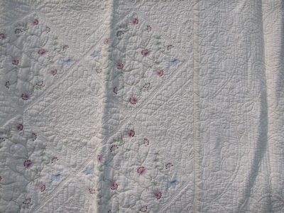 The Fairytale Flower Garden Quilt is a textured Ivory quilt adorned with intricate embroidery designs showcasing red and pink flowers, green leaves, and subtle blue butterfly accents in diamond patterns.