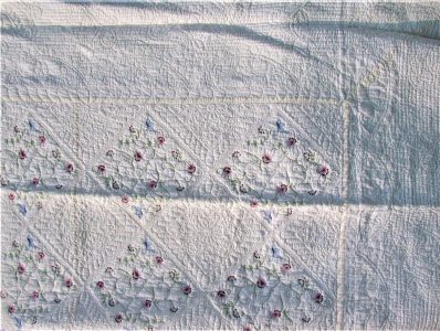 The Fairytale Flower Garden Quilt features light-colored cotton embroidery with intricate stitching patterns and floral designs showcasing pink and purple flowers. Its design incorporates geometric shapes and a textured surface.