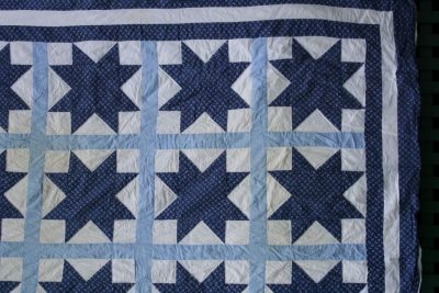 The Blue Paisley Morning Star Quilt showcases a star pattern in blue and white hues, featuring stars on a light blue and white grid background with dark blue dotted fabric as the backdrop within each star.