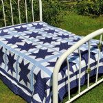 A white metal bed frame sits on a grassy lawn, adorned with the Blue Paisley Morning Star Quilt, featuring large blue stars and squares on a white and light blue background. Lush green plants and a stone wall surround the bed.