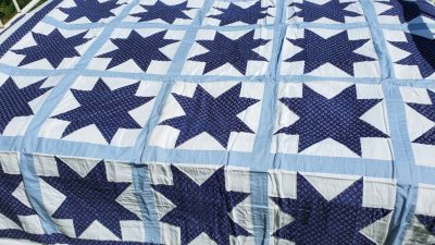 The Blue Paisley Morning Star Quilt showcases dark blue stars on a pale blue and white background, arranged in rows to create a geometric design, highlighting its intricate stitching and fabric details.