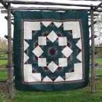 The Nostalgia 1980s Donna Sharp Broken Star Original Quilt with a multicolored geometric star pattern featuring dark green, ivory, and maroon colors is displayed on a rustic wooden frame against grass and trees.