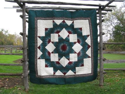The Nostalgia 1980s Donna Sharp Broken Star Original Quilt with a multicolored geometric star pattern featuring dark green, ivory, and maroon colors is displayed on a rustic wooden frame against grass and trees.