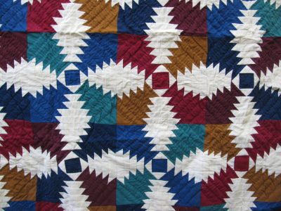 The Flannel Pineapple Hand Quilted 100% Cotton-Jewel Tones quilt features a geometric patchwork of interlocking diamonds in vibrant shades of blue, red, teal, ivory, and brown.