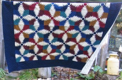 The Flannel Pineapple Hand Quilted 100% Cotton-Jewel Tones, featuring a vibrant geometric pattern in red, blue, gold, and ivory with interlocking shapes creating a colorful mosaic, hangs outdoors. A milk can and some plants are visible in the background.
