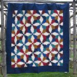 The Flannel Pineapple Hand Quilted 100% Cotton-Jewel Tones quilt, showcasing intricate geometric patterns in red, blue, brown, and ivory shapes resembling pine cones on a deep blue border, hangs on a wooden frame outdoors over grass.