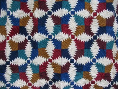 The Flannel Pineapple Hand Quilted 100% Cotton-Jewel Tones quilt features a geometric pattern with zigzag shapes in blue, red, brown, ivory, creating a vibrant and intricate design with repeating motifs.