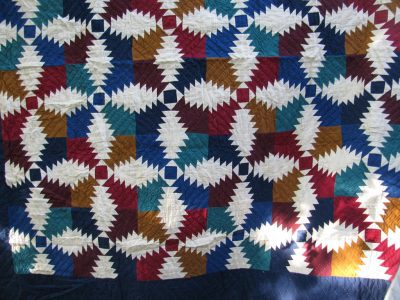 A quilt with a complex geometric pattern featuring ivory star-like shapes on red, blue, green, brown, and yellow fabrics arranged in a repeating pattern is available as the Flannel Pineapple Hand Quilted 100% Cotton-Jewel Tones.
