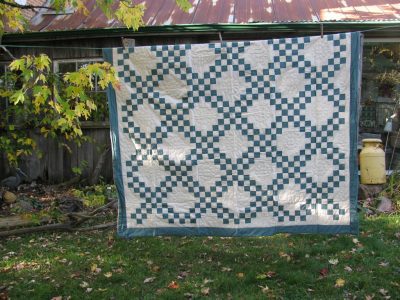 A Double Irish Chain Handmade Quilt in Antique Green with geometric designs hangs outside on a line. In the background, an old wooden shed with a tin roof is surrounded by scattered autumn leaves.