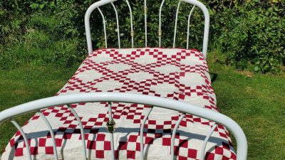 An Irish Chain Quilt- Burgundy rests on a white metal bed frame outdoors on a grassy lawn, featuring intricate red and white checkered patterns. Lush green foliage surrounds the scene in the background.