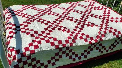 An Irish Chain Quilt- Burgundy with a red and white checkerboard pattern and floral-like designs is draped over a white metal bed frame, placed on a grassy lawn under sunlight.