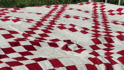 A close-up of the Irish Chain Quilt- Burgundy, showcasing a red and white checkerboard with diamond shapes. Spread out in sunlight, it highlights the fabric's texture against a grass backdrop.