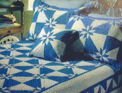 The bed features a Denim Star Hand-Quilted Quilt made of 100% cotton, showcasing a blue and ivory geometric pattern with star shapes. Matching pillows adorn the headrest, and a wooden nightstand with books sits beside the bed.