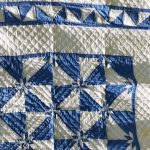 The Denim Star Hand-Quilted Quilt, made of 100% cotton, features a geometric pattern with blue and ivory triangles, squares, and diamond shapes, displayed on a grassy surface.