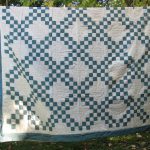 The Double Irish Chain Handmade Quilt - Antique Green is elegantly displayed outdoors, showcasing an intricate pattern of interlocking teal squares on a ivory background. It hangs from a line, amidst lush green foliage with a wooden chair in the backdrop.