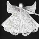 A Tree Ornament Angel made of Battenburg lace with intricate patterns, featuring wings and a ribbon around the neck. The solid black background highlights the delicate lace details.