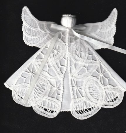 A Tree Ornament Angel made of Battenburg lace with intricate patterns, featuring wings and a ribbon around the neck. The solid black background highlights the delicate lace details.