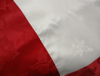 Close-up of the Christmas Red & Silky Ivory Jacquard Damask Tablecloth, showcasing a red section with a subtle pattern of holly leaves and berries, and a silky ivory section adorned with faint festive symbols such as stars and a bell with a ribbon.