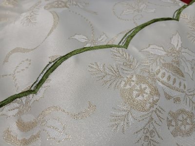A close-up of the Sparkle Christmas Motifs Green and Golden Tablecloth reveals sparkle ivory fabric adorned with golden embroidered holiday motifs, such as holly leaves and ornaments, along with a decorative green wavy seam as the finishing edge.