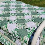 Set outdoors on grass, the bed features the Sparkling Green & White Courthouse Steps Prairie Point Border quilt with floral and geometric patterns. The bed has a white metal frame adorned with brass accents.