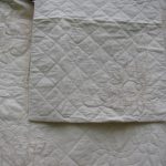 Close-up of the Ivory Pearl Braid Quilt, featuring beige quilted fabric with intricate floral embroidery. Spiral and leaf motifs create an elegant, textured appearance. A folded sham in the center highlights these details, emphasized by the scallop piping edge.