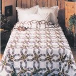 A bed with a Purple & Green 1980s Double Wedding Ring Quilt on an ornate metal headboard, set against wooden paneled walls. The room includes white pillows, a wooden cabinet, and decorative plants, welcoming ambiance of rustic charm