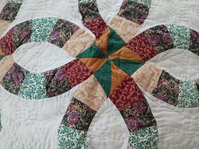 A close-up of the "Purple & Green 1980s Double Wedding Ring Hand Quilting" quilt showcases a multicolored pattern with interlocking rings, featuring floral and abstract designs in green, red, gold, and beige on a white quilted background.