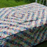 The Broken Dish Rare Trip Around the World Quilt, showcasing a multicolored geometric design with blue, green, pink, and white triangles forming diamonds, rests on a white metal bed frame outdoors. Grass surrounds the setup.