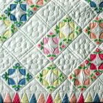 The Sunshine Cathedral Window Ice Cream Colours Hand-Quilted Quilt showcases a vibrant, geometric design on a white background with diamond patterns in blue, green, pink, and orange. The quilt's edges are adorned with pink and blue triangular shapes featuring intricate quilting and floral motifs.