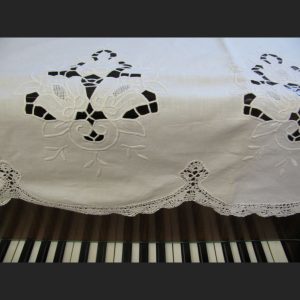 The Cluny Lace Piano Cover Scarf with Richelieu Embroidery (90"x30") drapes over a piano’s top edge, its white embroidered floral design partially covering the black and white keys.