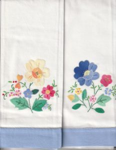 Set of 2 white Hand-Stitched Appliqué Guest Towels, each with a hemstitch pastel border and colorful embroidered flowers—yellow, pink, red, and blue—and green leaves in various arrangements.